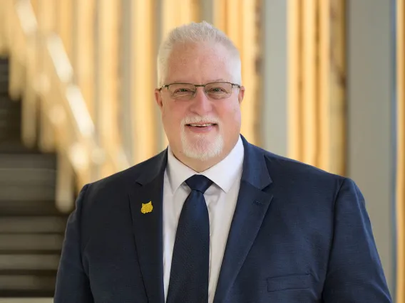 White man with grey hair, mustache and beard stands smiling. He is wearing a navy blue suite and tie with a white shirt. He is wearing a University or Arizona Wildcat pin on his lapel.