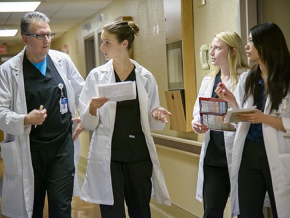 Female white coat students walking down hallway in discussion with male physician faculty.
